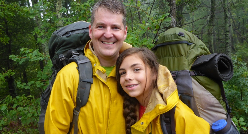 A parent and child pose for a photo in a green wooded area. Both are wearing rain jackets and backpacks.